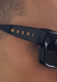 Occhiali da sole neri con montatura spessa, caratterizzati da un evidente lettering giallo e rosso "MARNI" sulle tempie. Texture liscia e lucida.