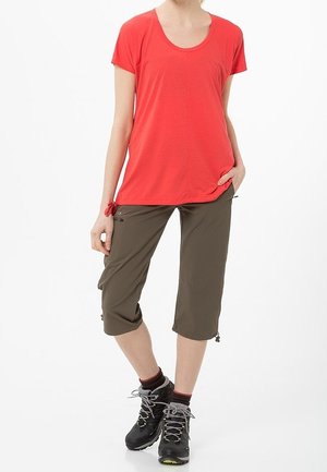 T-shirt rossa a maniche corte con scollo a barca, abbinata a pantaloni verde oliva fin sopra il ginocchio con coulisse e zip. Stivali da trekking neri.