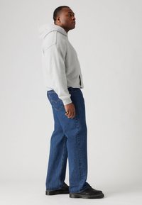 Gray hooded sweatshirt with a front pocket, paired with blue denim jeans. Individual stands sideways, wearing black platform shoes.