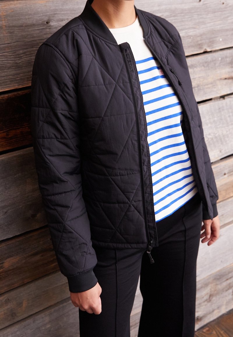 Svart vadderad bomberjacka med ribbade ärmar och krage, med en framfickzipper. Under jackan en vit skjorta med blåa horisontella ränder.