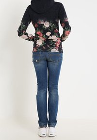 Sudadera con capucha en azul oscuro con estampado floral en la parte inferior, combinada con jeans ajustados azules y zapatillas blancas.