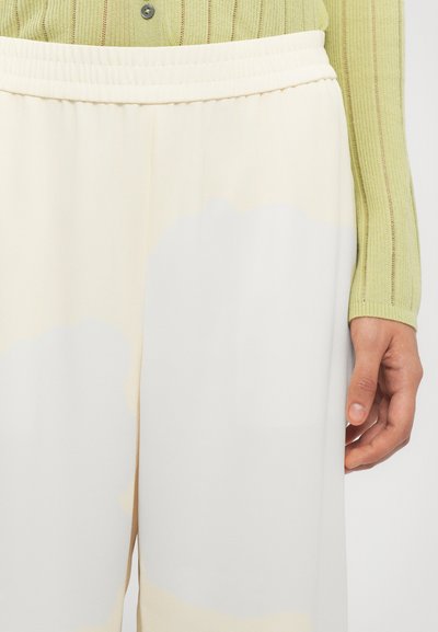 Fließende beigefarbene Hosen mit sanftem Glanz und einem weißen abstrakten Muster. Ein elastischer Bund und ein strukturiertes grünes Strickoberteil sind sichtbar.