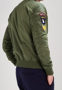 Giacca bomber verde oliva con polsini e orlo a coste, caratterizzata da toppe ricamate sulla manica sinistra, che mette in evidenza dettagli ispirati all'ambiente militare.