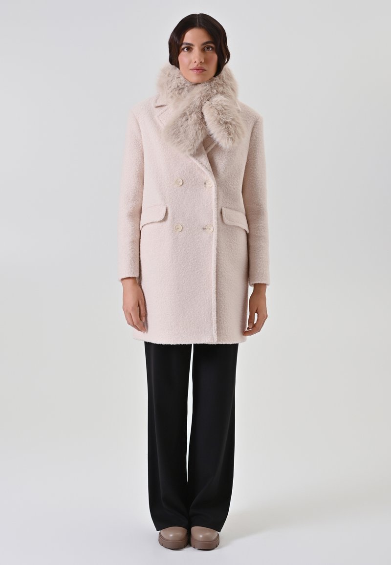 Manteau double boutonnage rose clair avec un col duveteux, deux poches avant, tissu texturé, associé à un pantalon noir et des chaussures beiges.