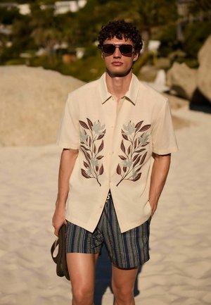 Young man wearing sunglasses, a beige short-sleeve shirt with leaf patterns, and striped shorts, walking on a sandy beach holding sandals.