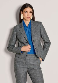 Grijs geruite blazer met twee knopen, gecombineerd met een blauwe gedrapeerde top. Het model draagt grote crème oorbellen, die het gestructureerde ontwerp van de outfit benadrukken.