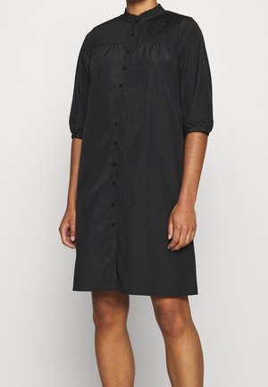 Femme portant une robe noire boutonnée au genou, manches trois-quarts et détail plissé sur le plastron.