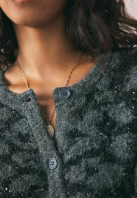 Sivi pleteni kardigan s teksturiranom, pahuljastom površinom. Ima dva tamna gumba i suptilne perle kao ukras na tkanini.