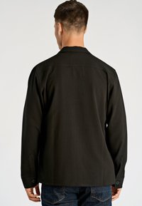 Zwarte lange mouwen blouse met een spitse kraag, gestructureerde stof en afgewerkte manchetten met knopen. Achteraan zicht op het eenvoudige ontwerp en de getailleerde pasvorm.