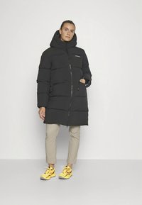 Juoda pūkinė striukė su gaubtu, su užtrauktuku ir dviem šoninėmis kišenėmis, dėvima ant smėlio spalvos kelnių ir geltonų sportbačių.