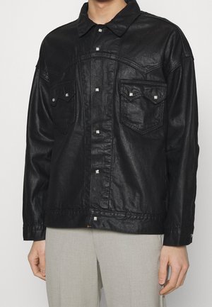 Veste noire en cuir synthétique avec fermeture à boutons et poches poitrine à rabat portée par une personne avec un pantalon gris clair sur un fond uni.
