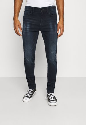 Temno modri jeans z ožjim krojem, obrabljenimi detajli in klasičnim pet-pocket dizajnom, v kombinaciji s črnimi in belimi športnimi copati.