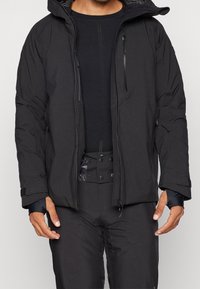 Muž v černé venkovní bundě s kapsou na zip na hrudi, černém dlouhém rukávu a černých kalhotách s knoflíkovým páskem.