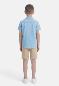 Αγόρι που στέκεται με την πλάτη, φοράει ανοιχτό μπλε κοντομάνικο πουκάμισο, μπεζ σορτς και λευκά αθλητικά παπούτσια, με φόντο απλό λευκό.