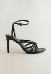 Schwarze High-Heel-Sandale mit geflochtenen Riemen und geknotetem Design. Der Schuh hat eine glatte Ledersohle und einen verstellbaren Knöchelriemen.