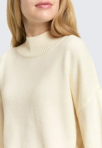 Kremowy dzianinowy sweter z wysokim, prążkowanym kołnierzem i luźnymi rękawami, charakteryzujący się miękką teksturą i minimalistycznym wykończeniem.