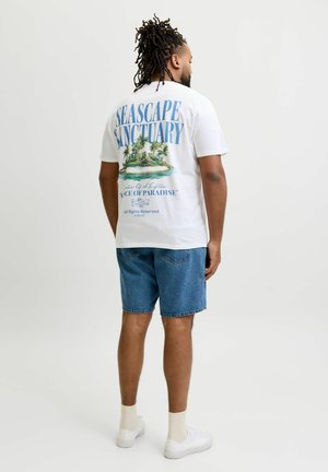 Mann mit Dreadlocks trägt ein weißes T-Shirt mit "Seascape Sanctuary" und Inselgrafik auf dem Rücken, blaue Jeansshorts, weiße Socken und weiße Sneakers.