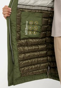 Groene geïsoleerde jas met een gevoerde binnenkant, voorzien van een waterdichte membraanlabel en zichtbare ritsen. Gelaste naden voor duurzaamheid.