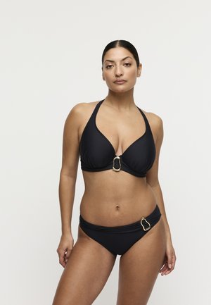 Vrouw staand, gekleed in een zwarte halterbikini met ovale ringen in goudkleur op de top en de onderkant, tegen een effen witte achtergrond.