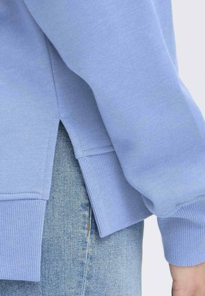 Lyseblå sweater med ribbestrikkede manchetter og kant, med en slidse i siden for ekstra detalje, lagt over blå denimstof.