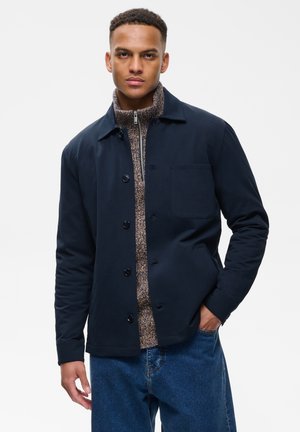 Homme portant une veste bleu marine à boutons par-dessus un pull marron zippé et un jean bleu, debout avec une main dans la poche sur fond blanc.