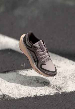 Grauer und beiger Sportschuh mit schwarzen Akzenten und reflektierenden Details, schwebt über einer schwarzen Asphaltfläche mit weißen Straßenmarkierungen.