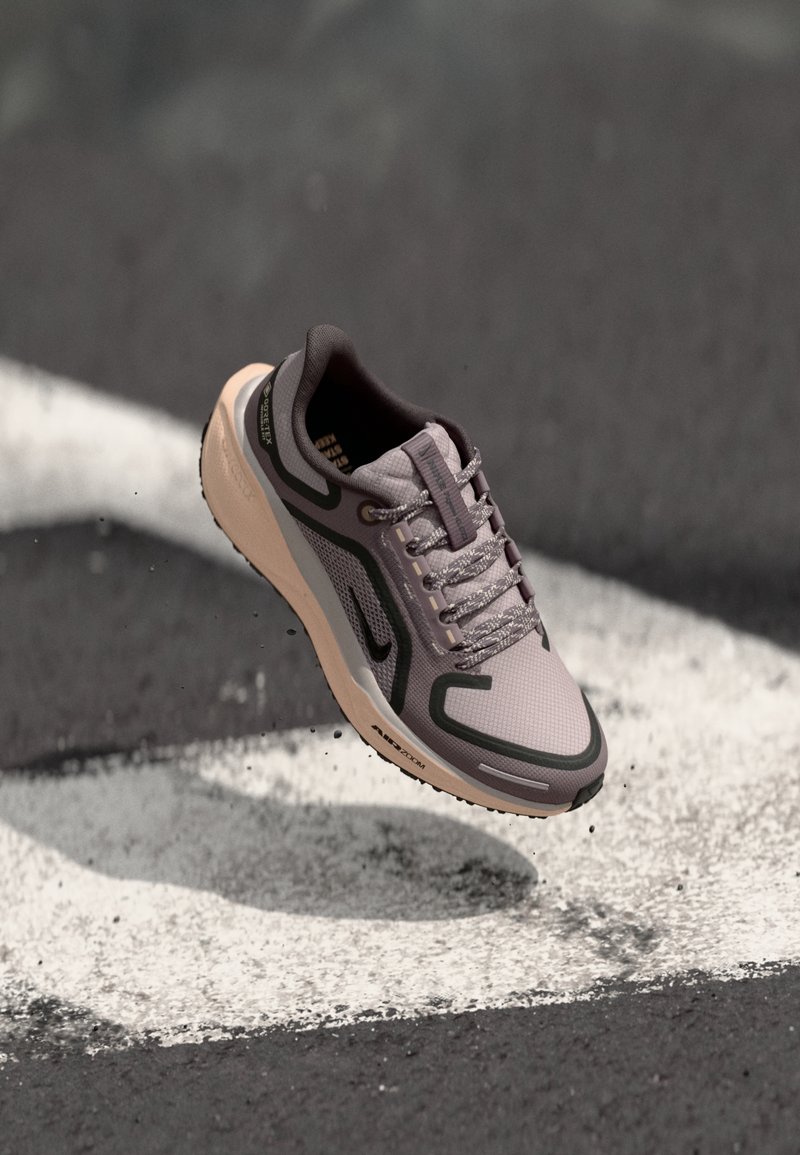 Chaussure de sport grise et beige avec des accents noirs et des détails réfléchissants, en suspension au-dessus d'une surface d'asphalte noire avec des marquages routiers blancs.