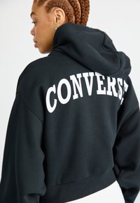 Black cropped hoodie made of cotton. Features a large white "CONVERSE" logo on the back. Soft texture with a hood and relaxed fit.