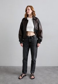 Giacca bomber in pelle marrone, top corto bianco, jeans grigio scuro e sandali neri con cinturini. Trame piatte e lisce con hardware minimal.