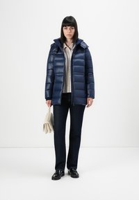 Kvinne iført en marineblå pufferjakke, stripete skjorte, mørke jeans, svarte sko, og holder en beige håndveske foran en enkel hvit bakgrunn.