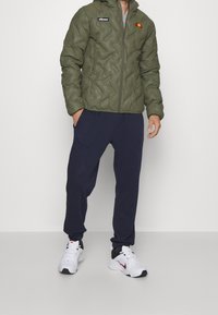 Mand iført en olivengrøn quiltet jakke, navy joggers og hvide Nike sneakers, stående mod en ensfarvet baggrund.