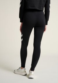 Leggings noirs en tissu extensible, avec une texture lisse et des rayures blanches diagonales sur les côtés. Portés avec des chaussures de sport légères.