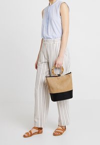 Borsa tote intrecciata beige con base nera e manici in stile bambù. Abbinata a una blusa senza maniche azzurro chiaro e pantaloni a righe.