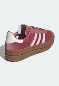 Tenisky Adidas Gazelle ve tmavě růžové semišové úpravě s světle růžovými pruhy, bílým podpatkem a silnou hnědou gumovou podrážkou. Texturovaný svršek s akcenty značky.