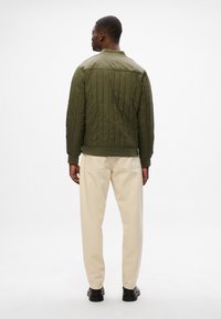 Giacca bomber trapuntata verde oliva con colletto e polsini a costine, indossata con pantaloni chiari e scarpe nere, vista da dietro.