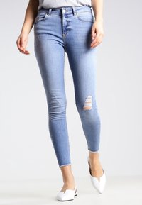 Jeans ajustados de color azul claro hechos de mezclilla, con una rodilla desgastada y bordes deshilachados, que presentan una silueta entallada. Combinados con zapatos blancos de slip-on.