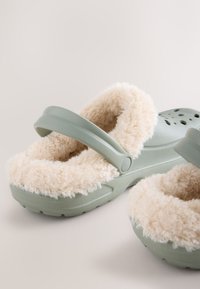 Gröna gummiklumpor med en mjuk, krämfärgad fuskpälsfoder. Har en bred rem över toppen och ventilationshål på sidorna.