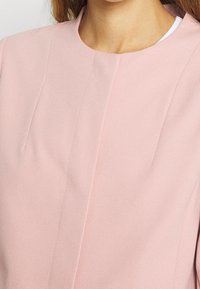 Blazer de color rosa claro con escote redondo, tejido sutilmente texturizado y detalles de costura minimalistas. Presenta un ajuste limpio y entallado.