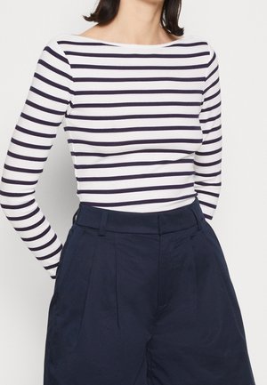 Woman wearing a long-sleeve white top with navy horizontal stripes tucked into high-waisted navy pleated pants.