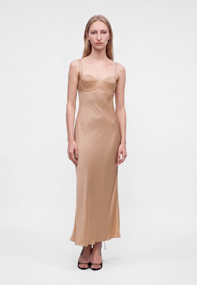 Femme aux longs cheveux blonds portant une robe beige à fines bretelles et des talons ouverts noirs, debout devant un fond blanc uni.