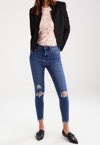 Musta bleiseri vaaleanpunaisen pitsitopin päällä, yhdistettynä kuluneisiin sinisiin skinny-farkkuihin, joissa on repaleiset helmavedokset ja reijät polvissa. Mustat kengät.