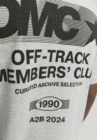 Camiseta de algodón gris con texto en negrita negro que dice "CLUB DE MIEMBROS FUERA DE RUTA" y elementos gráficos. Incluye detalles de "1990" y "A2B 2024".