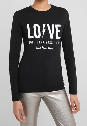 Osoba nosząca czarną koszulę z długim rękawem z białym napisem "LOVE" z błyskawicą i słowami "JOY • HAPPINESS • FUN" nad srebrnymi metalicznymi spodniami.