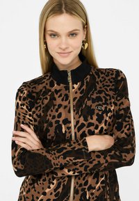 Giacca leopardata in marrone e nero con zip frontale, colletto nero e dettaglio logo dorato sul petto, realizzata in un tessuto liscio.