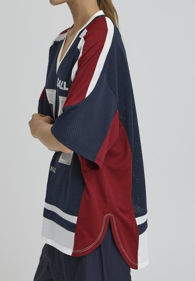 Personne portant un maillot de sport ample de couleur marine, rouge et blanche en tissu perforé avec le numéro 77, se tenant les bras croisés.