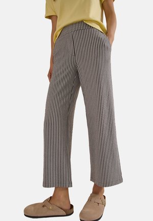 Person iført grå og hvide lodrette stribede bukser med vide ben kombineret med beige slip-on sko og en gul top.