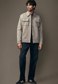 Beige tekstureret jakke med to forlommer, knaplukning, båret over en sort skjorte. Sammen med mørke jeans og sorte sneakers.