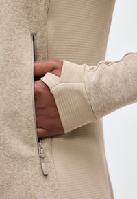 Tamsios odos ranka su dėvimu smėlio spalvos ribbingu rankovėle įkišta į smėlio spalvos užtraukiamą kišenę ant tekstūruoto audinio drabužio.