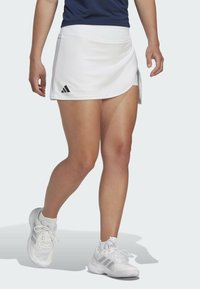 Balta sporta svārku ar kārtotu apakšmalu, sānu izgriezumu un melnu Adidas logo. Valkāta ar baltām sporta kurpēm ar pelēkiem akcentiem.