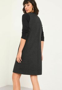 Schwarzes Kleid aus strukturiertem Stoff, mit langen schwarzen Ärmeln und Knopfdetails auf dem Rücken. A-Linien-Silhouette mit knieumspielendem Schnitt.